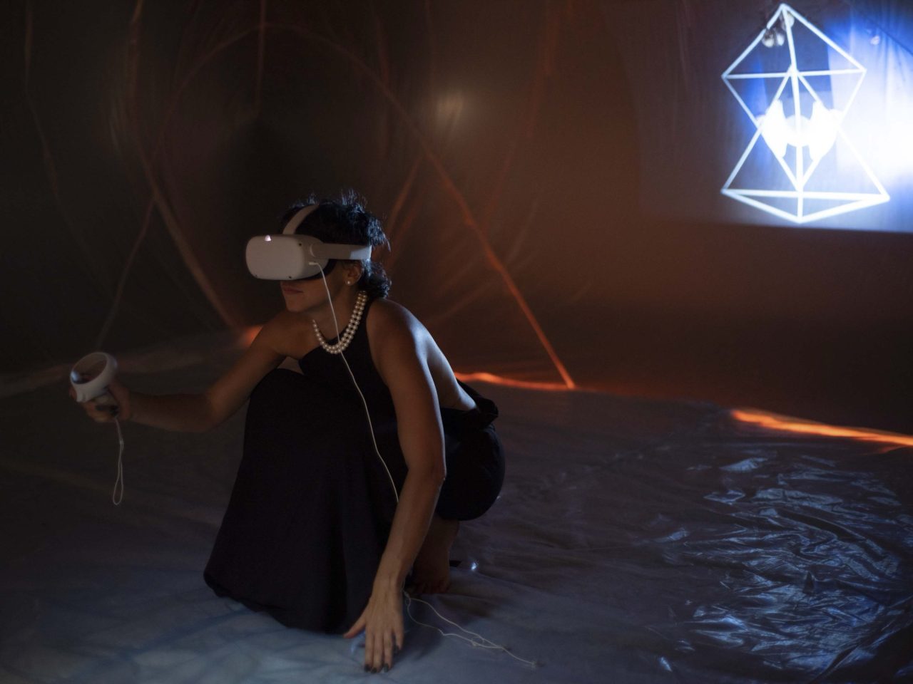 A person kneeling in a darkened room wearing a VR headset, a luminous wireframe form projected on the wall behind them.