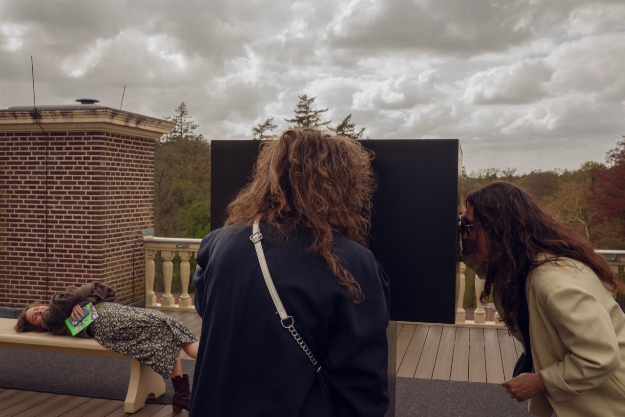 Visitors on the roof terrace of Het Loo Palace looking into a large black diorama box under an overcast sky.